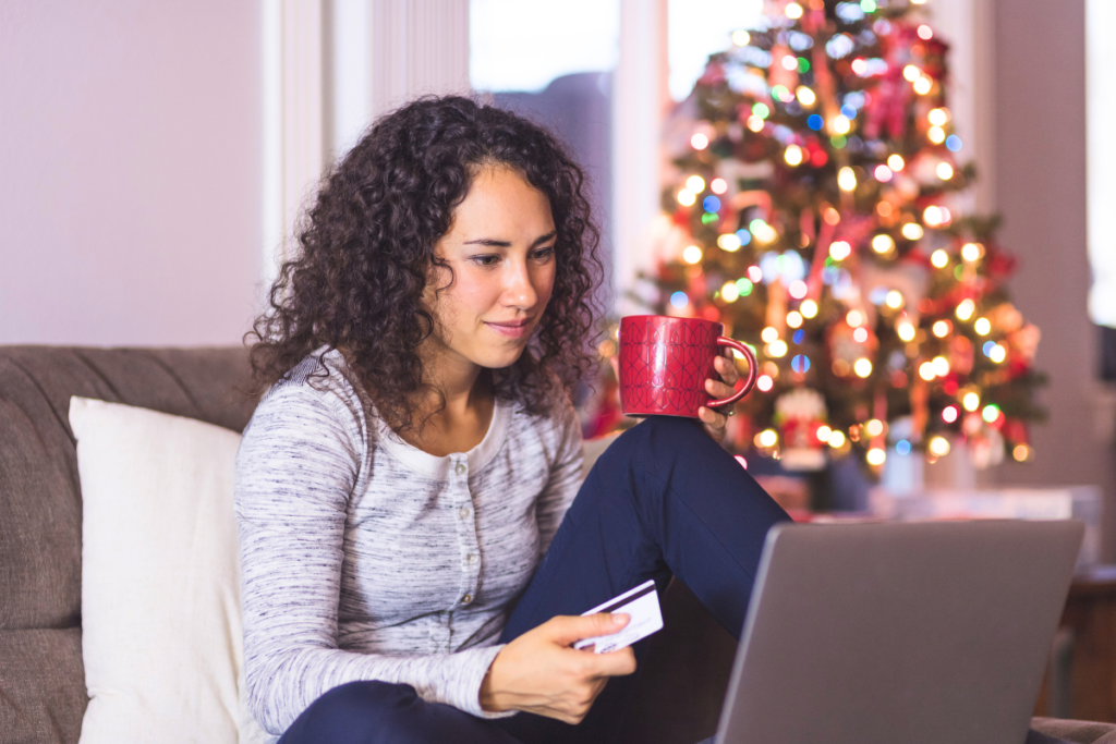 Una chica tomandose una taza de algo caliente mientras está al lado del arbol de navidad realizando una compra online en la web de mikamiseta